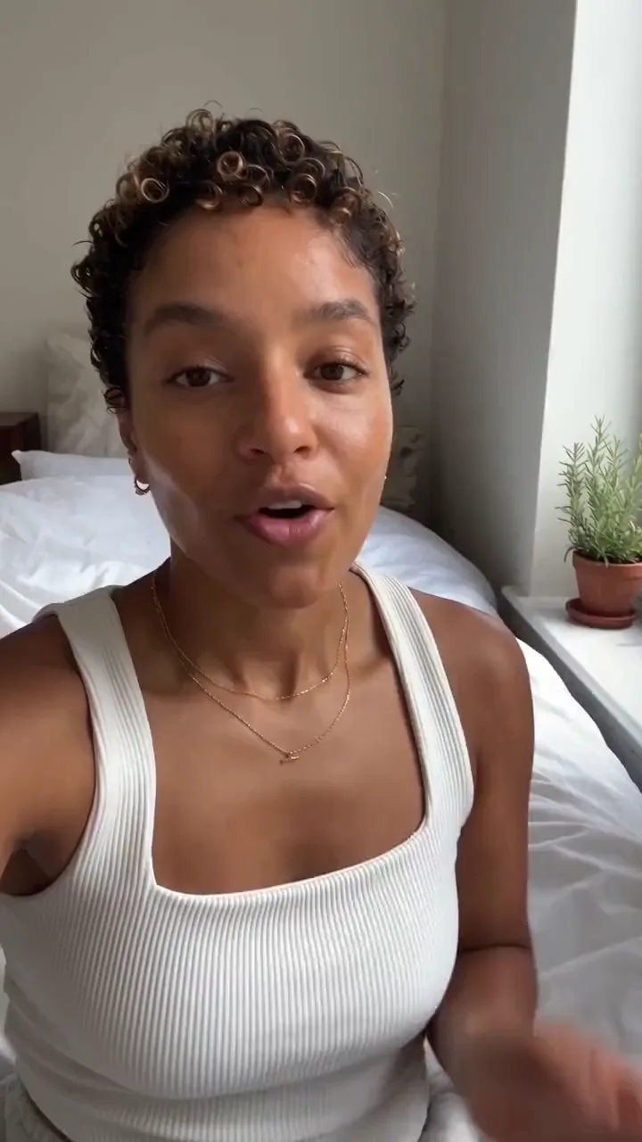 A creator on her bed talks through a tinted moisturizer and dabs it on one cheek